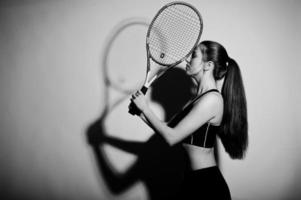 retrato preto e branco do jogador de mulher jovem e bonita em roupas esportivas, segurando a raquete de tênis em pé contra um fundo branco. foto