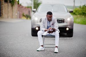 homem afro-americano rico e elegante de blazer e calça branca, óculos com revista sentado na cadeira contra o carro suv. foto