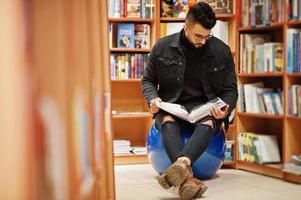 homem alto estudante árabe inteligente, use jaqueta jeans preta e óculos, sentado na biblioteca e leia o livro nas mãos. foto