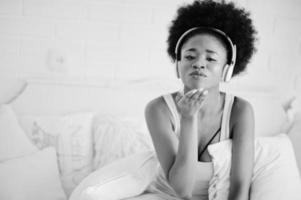 jovem afro-americana sentada na cama e ouvir música em fones de ouvido. foto