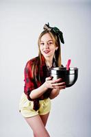 jovem dona de casa engraçada na camisa quadriculada e short amarelo pin up estilo com panela e colher de cozinha isolado no fundo branco. foto