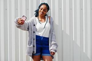 mulher africana com cabelo dreads, em shorts jeans posou contra a parede de aço branca e fazendo selfie por telefone. foto