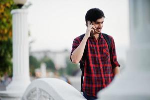 homem jovem estudante indiano na camisa quadriculada e jeans com mochila falando no celular. foto