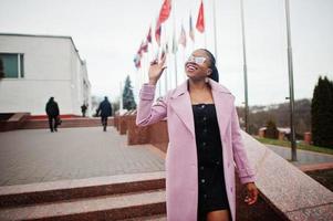 jovem elegante linda afro-americana na rua, vestindo óculos e casaco de roupa de moda, contra bandeiras de diferentes países do mundo. foto