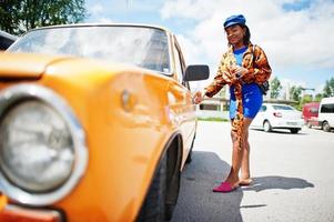 bela dama afro-americana com óculos de sol em pé perto de carro retrô clássico laranja. foto
