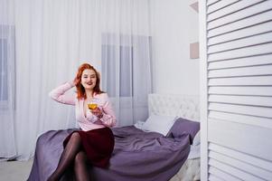 linda garota ruiva de blusa rosa e saia vermelha com copo de vinho na mão na cama no quarto. foto