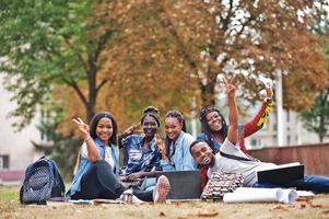 grupo de cinco estudantes universitários africanos passando tempo juntos no campus no pátio da universidade. amigos negros afro sentados na grama e estudando com laptops. foto