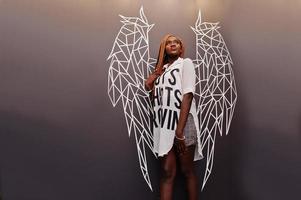 linda mulher africana em roupas casuais elegantes posando contra a parede de asas de anjo.