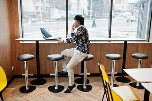 jovem freelancer indiano masculino sentado computador portátil aberto no café de fast food, bonito homem asiático falando telefone durante o trabalho no net-book no café confortável. foto