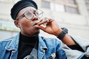 feche o retrato do homem afro-americano na jaqueta jeans, boina e óculos, fumando charuto. foto