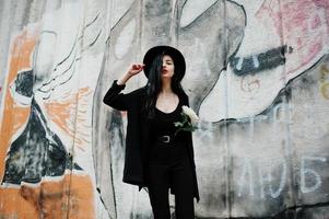 garota sensual toda em preto, lábios vermelhos e chapéu. mulher dramática gótica segura flor de crisântemo branco contra parede de graffiti. foto