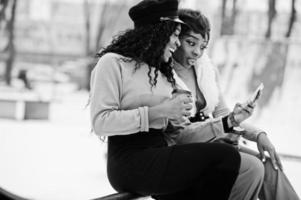 duas mulheres afro-americanas posaram no dia de inverno contra um fundo nevado com xícaras de café e olhando no telefone. foto