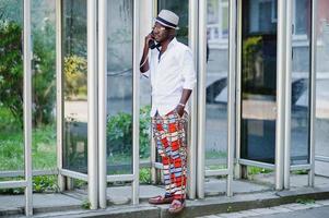 homem afro-americano elegante de camisa branca e calça colorida com chapéu e óculos posou ao ar livre na cabine telefônica. menino modelo preto na moda. foto