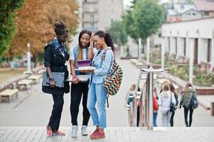 três estudantes africanos femininos posaram com mochilas e itens escolares no pátio da universidade e olham para o tablet. foto