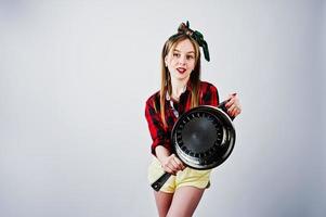 jovem dona de casa engraçada na camisa quadriculada e short amarelo pin up estilo com frigideira isolada no fundo branco. foto