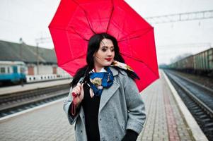 menina morena de casaco cinza com guarda-chuva vermelho na estação ferroviária. foto