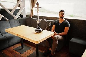 homem árabe de barba elegante de óculos e camiseta preta fumando cachimbo de água no bar da rua. modelo árabe descansando. foto