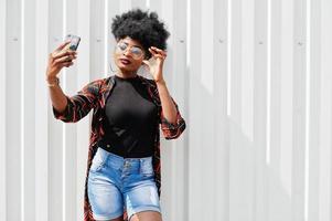 mulher africana com cabelo afro, em shorts jeans e óculos posou contra a parede de aço branca e fazendo selfie por telefone. foto