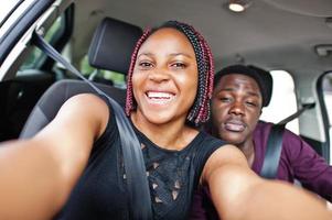 lindo jovem casal americano africano sentado nos bancos do passageiro da frente enquanto homem bonito dirigindo um carro. fazendo selfie juntos. foto