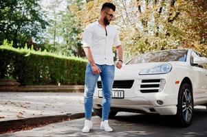 elegante modelo de homem árabe alto na camisa branca, jeans e óculos de sol posou na rua da cidade. barba cara árabe atraente rico contra carro suv branco. foto