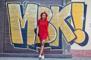 linda e magra garota afro-americana de vestido vermelho com dreadlocks e mochila posou ao ar livre na parede de grafite de fundo de rua. elegante modelo preto. foto