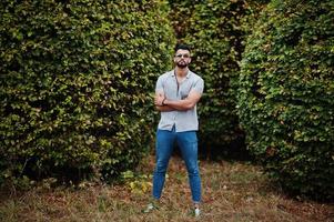 homem de barba árabe alto na moda usar camisa, jeans e óculos de sol posou no parque contra vegetação. foto