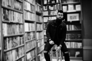 homem alto estudante árabe inteligente, use jaqueta jeans preta e óculos, na biblioteca sentado na cadeira contra prateleiras de livros. foto