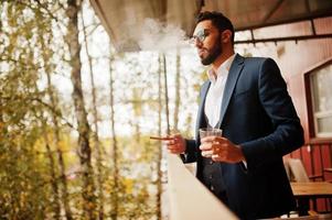 bonito homem árabe bem vestido fuma charuto na varanda do pub. foto