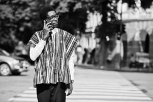 homem africano em roupas tradicionais e óculos andando na faixa de pedestres e falando no celular. foto