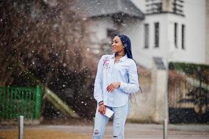 menina afro-americana elegante com dreads segurando o celular na mão, clima de neve ao ar livre. foto