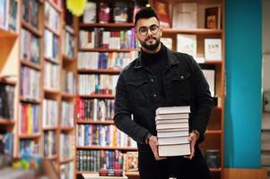 homem alto estudante árabe inteligente, use jaqueta jeans preta e óculos, na biblioteca com pilha de livros. foto