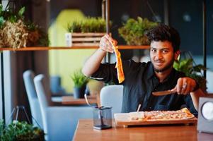 confiante jovem indiano na camisa preta, sentado na pizzaria com pizza. foto