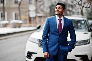 modelo de homem elegante indiano elegante terno posado em dia de inverno contra carro suv de negócios branco. foto