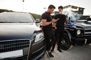 dois irmãos asiáticos usam todo preto posando perto de carros suv e fumam cachimbo de água. foto