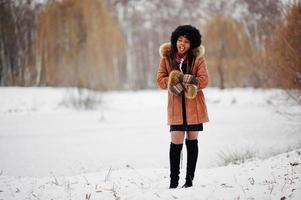 mulher afro-americana de cabelo encaracolado usa casaco de pele de carneiro e luvas posadas no dia de inverno, mostra que está frio. foto