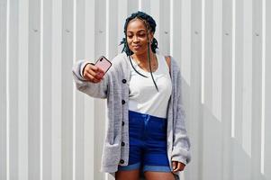 mulher africana com cabelo dreads, em shorts jeans posou contra a parede de aço branca e fazendo selfie por telefone. foto