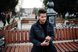homem indiano bonito e elegante na jaqueta jeans preta posou ao ar livre, sentado no banco. foto