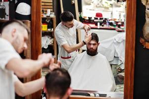 jovem barbudo cortando o cabelo pelo cabeleireiro enquanto está sentado na cadeira na barbearia. alma de barbeiro. foto