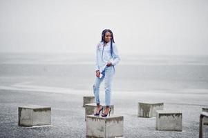 menina afro-americana elegante com dreads segurando o celular na mão, ao ar livre no cais contra o lago congelado em tempo de neve. foto