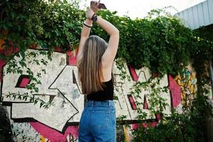 Garota de hipster casual elegante de boné, óculos e calça jeans, ouvindo música de fones de ouvido do celular contra a grande parede de graffiti. foto