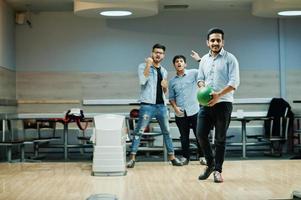 homem do sul da Ásia em pé de camisa jeans na pista de boliche com bola nas mãos. cara está se preparando para um lance. amigos o apoiam ruidosamente. foto