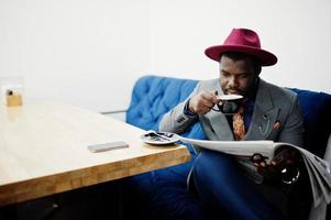 modelo de homem afro-americano elegante em gravata cinza e chapéu vermelho bebe café no café e lê jornais. foto