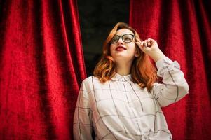 mulher ruiva atraente em óculos, use blusa branca posando no arco de cortinas vermelhas abertas. foto