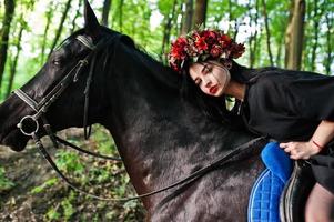 garota mística em grinalda veste de preto a cavalo em madeira. foto