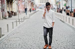 homem elegante modelo indiano em roupas casuais e óculos de sol posou ao ar livre na rua da índia e falando no celular. foto