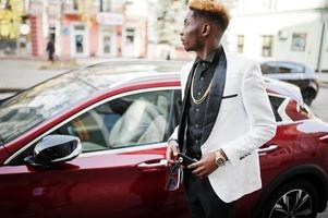 homem afro-americano bonito chique de terno branco contra carro de luxo vermelho com telefone celular na mão. foto