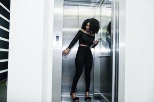 elegante jovem e bela mulher de negócios afro-americana com penteado afro vestir em preto elegante, ficar no elevador com telefone e laptop nas mãos. foto