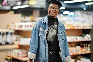 homem afro-americano casual elegante na jaqueta jeans e boina preta segurando duas cestas, andando e fazendo compras no supermercado. foto