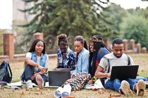 grupo de cinco estudantes universitários africanos passando tempo juntos no campus no pátio da universidade. amigos negros afro sentados na grama e estudando com laptops. foto