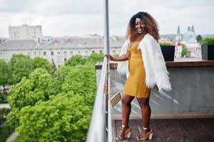 glamour mulher afro-americana de vestido amarelo e capa de lã branca com bolsa posada na varanda. foto